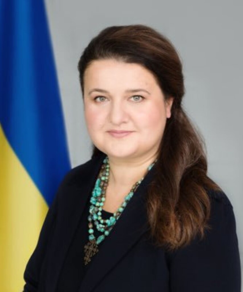 A white woman standing in front of the Ukranian flag which is yellow and blue. 