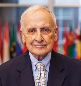 Headshot of white older male with dark suite and yellow and blue patterned tie.