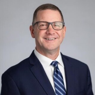 Headshot of white male with glasses wearing a dark suit and blue striped tie.